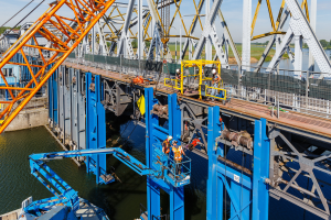 Eine Gruppe von Männern in Sicherheitswesten und Helmen arbeitet an der Konstruktion einer Eisenstabbrücke über einen Gewässser, umgeben von einem Zaun, Bäumen, Gebäuden und einem klaren blauen Himmel.