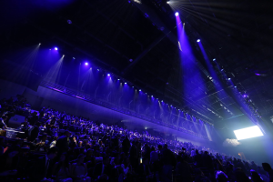 Eine große Menschenmenge sitzt einer Bühne mit Deckenbeleuchtung gegenüber, und auf der rechten Seite ist ein Bildschirm zu sehen, was auf einen Veranstaltungsort hindeutet.