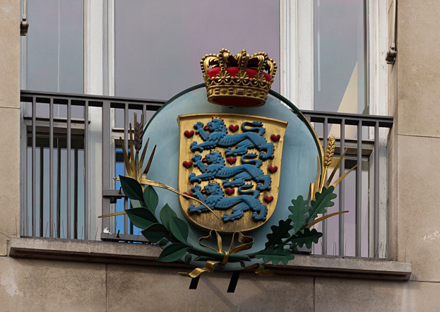 Das britische Königswappen auf der Fassade eines Gebäudes dargestellt, begleitet von einem Wappen mit Krone und Blattlogo, mit Fenstern und Sicherheitsgittern im Hintergrund.