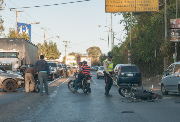 Gruppe von Menschen um ein verunglücktes Motorrad auf der Seite einer Straße mit mehreren Fahrzeugen, Bäumen, Polen, Lampen, Schildern und dem Himmel im Hintergrund.
