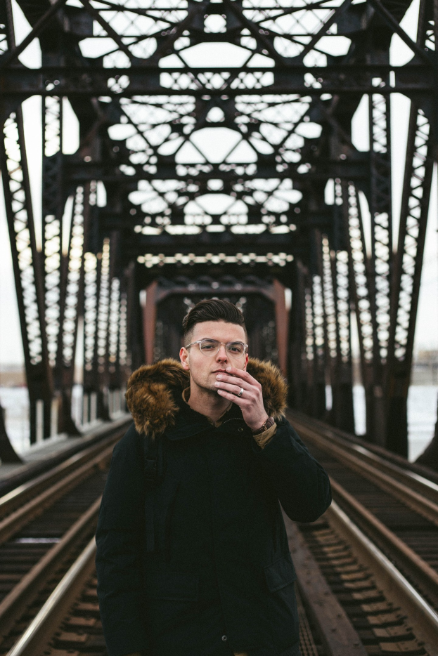 Ein Mann in einer Jacke und mit Brille steht auf einem Bahngleis und raucht eine Zigarette, im Hintergrund eine Brücke und der Himmel.