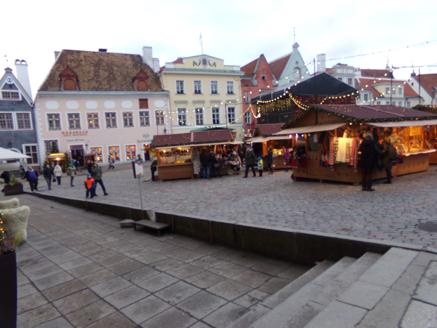 Ein belebter Weihnachtsmarkt in Tallinn, Estland mit Menschen um geschmückten St├Ąnden, festlicher Beleuchtung, bew├Âlktem Himmel, Treppen und Topfpflanzen im Bereich.