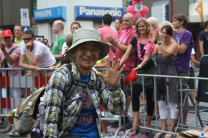 Ein Mann mit Hut, Sonnenbrille und Brille steht vor einer Menge bei einer Christopher Street Day Parade, mit einigen klatschenden Menschen und einer Absperrung dahinter und Gebäuden mit Namensschildern und Luftballons im Hintergrund.