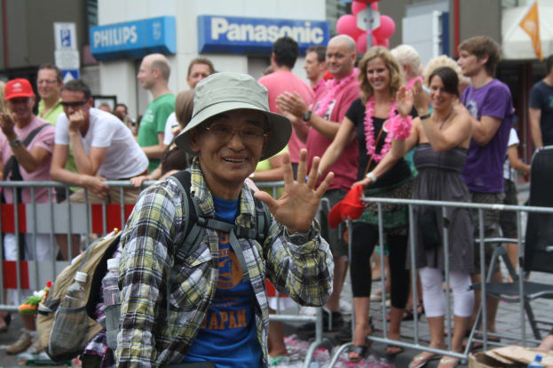 Ein Mann mit Hut, Sonnenbrille und Brille steht vor einer Menge bei einer Christopher Street Day Parade, mit einigen klatschenden Menschen und einer Absperrung dahinter und Gebäuden mit Namensschildern und Luftballons im Hintergrund.