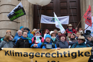 Eine Gruppe von Menschen steht vor einem Gebäude mit Säulen und einer Tür, hält ein Transparent mit Text, Fahnen mit Stangen und Luftballons und nimmt an einem Klimastreik in Deutschland teil.