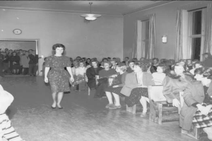Eine Frau in einem schwarzen und weißen Kleid geht über einen Laufsteg während einer Modenschau für Frauenwahlrecht, mit einer Menge, die auf Stühlen sitzt und einer Uhr und Lichtern an der umgebenden Wand.