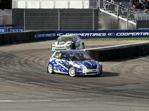 Zwei Autos auf einer Rennstrecke mit Maschendrahtzaun und Bannern, umgeben von Zuschauern auf einer Treppe mit Geländer, mit Bäumen und Zelten im Hintergrund auf einer grünen Wiese.