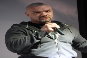 Ein Mann in einer schwarzen Kapuzenjacke und einem schwarzen Anzug mit einem weißen Hemd, der selbstbewusst vor einer Wand steht und offenbar ein Komiker ist.