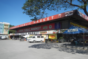 Öffentlicher Markt mit einem vor ihm geparkten Van, umgeben von Gebäuden, Geschäften, Namensschildern, Kraftfahrzeugen auf der Straße, Personen unter einem Zelt sitzend, Geländern, Bäumen und einem klaren blauen Himmel.