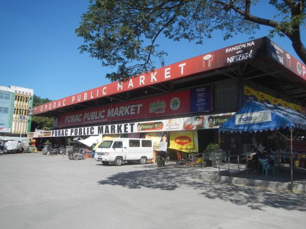 Öffentlicher Markt mit einem vor ihm geparkten Van, umgeben von Gebäuden, Geschäften, Namensschildern, Kraftfahrzeugen auf der Straße, Personen unter einem Zelt sitzend, Geländern, Bäumen und einem klaren blauen Himmel.