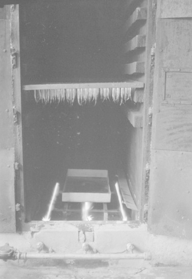 Schwarz-weiß-Foto eines Industrieofens mit einem brennenden Feuer in der Mitte, einer Tür an der linken Wand und Rohren, die am Boden entlanglaufen.