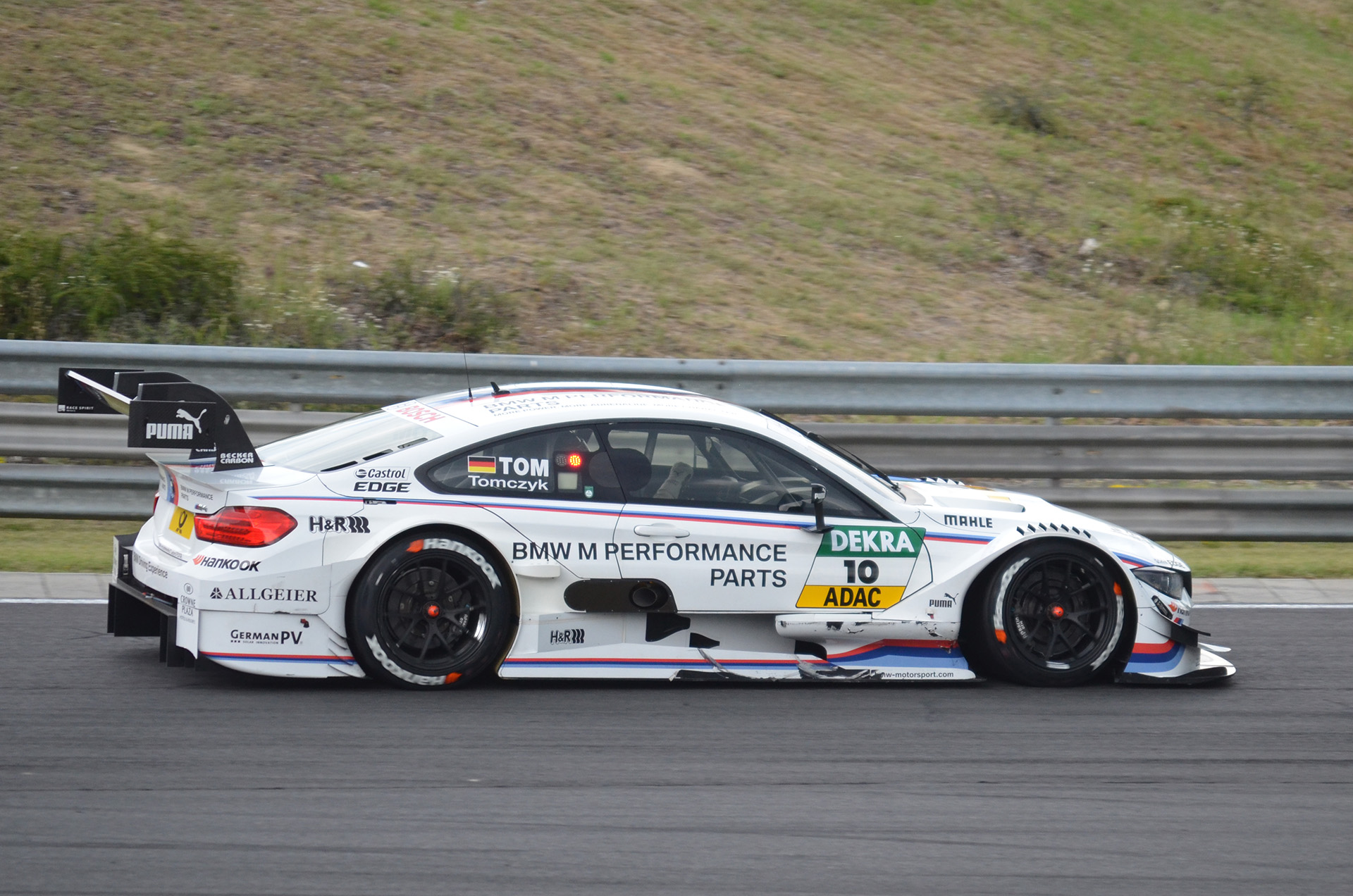 Ein BMW M4 DTM-Rennwagen fährt auf einer Rennstrecke, umgeben von grünem Gras, mit einer Absperrung im Hintergrund.