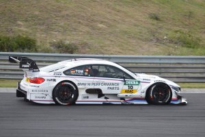 Ein BMW M4 DTM-Rennwagen fährt auf einer Rennstrecke, umgeben von grünem Gras, mit einer Absperrung im Hintergrund.