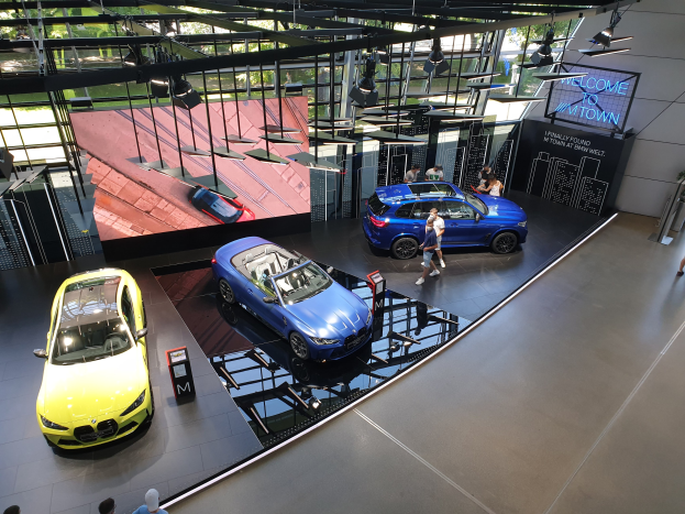 Eine Gruppe von Menschen, die im BMW Museum in München stehen, mit Autos auf der linken Seite und einem Schild mit der Aufschrift "BMW Museum München" auf der rechten Seite, mit Blick auf eine Outdoor-Szene von Büschen, Gebüuden und Himmel durch Glasfenster.