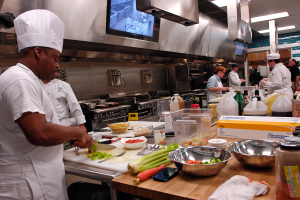 Eine Gruppe von Köchen in einer Küche bei der Zubereitung von Speisen auf einem Schneidbrett, wobei ein Koch im Vordergrund einen weißen Kittel und eine Haube trägt und ein Messer in der Hand hält und von verschiedenen Utensilien, Schüsseln, Gemüse, einem Mobiltelefon, Flaschen und anderen Gegenständen umgeben ist, mit einem Fernseher und Deckenleuchten im Hintergrund.