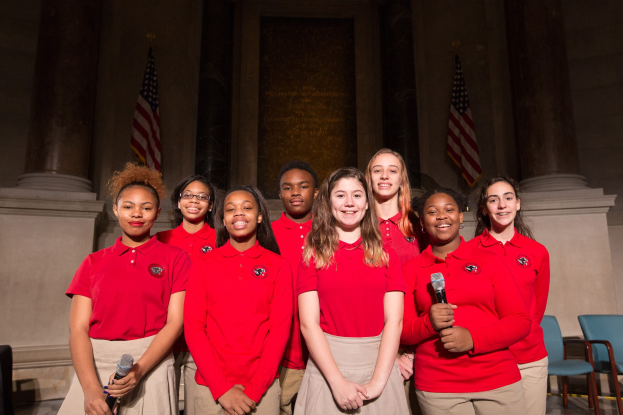 Gruppe junger Frauen in roten Hemden, die strahlend Mikrofone halten und vor Stühlen, Säulen, Fahnen und einer Wand stehen.
