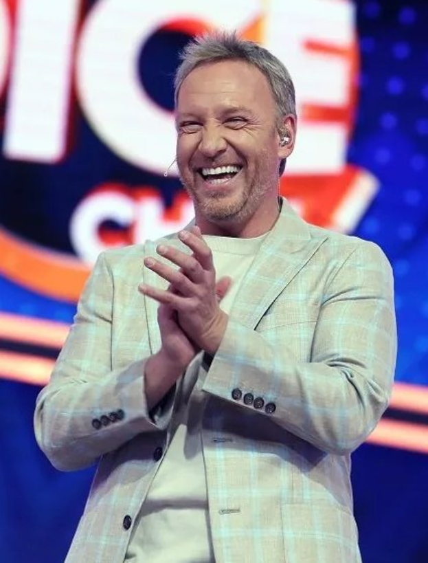 Ein lachender Mann in einem Anzug, der auf einer Bühne klatscht, mit 'The Voice'-Plakat im Hintergrund.