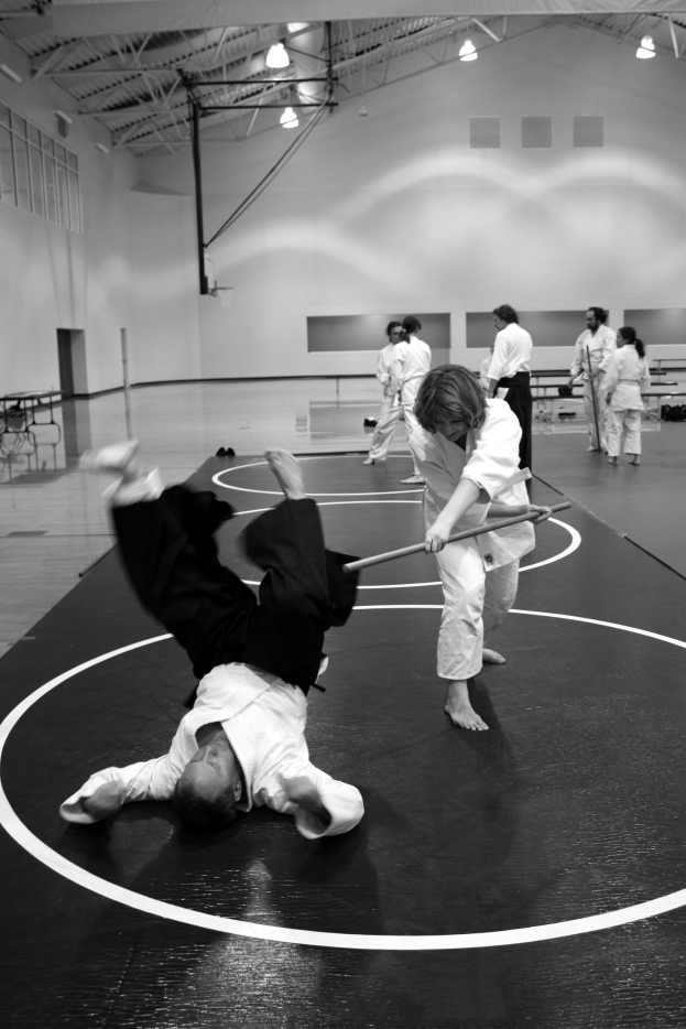 Zwei Personen üben Karate mit Stöcken in einer Turnhalle, während andere zusehen, mit sichtbaren Deckenlampen im Hintergrund.