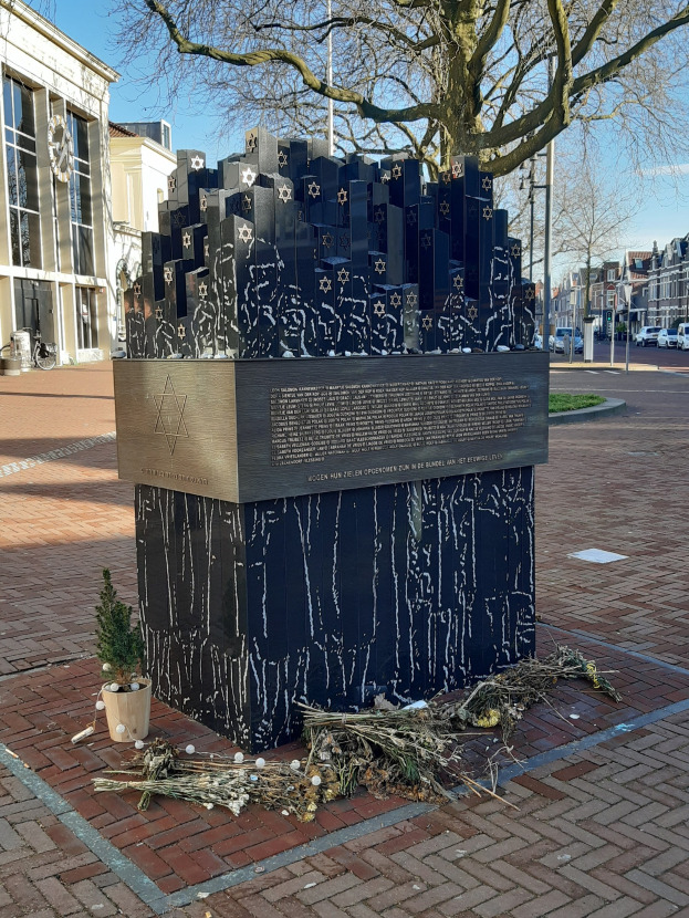 Denkmal in einem Stadtplatz für Holocaust-Opfer, mit einem Baum davor, einem Topf mit einer Pflanze, verstreuten trockenen Blättern und Gebäuden, Fahrzeugen, Polen und Bäumen im Hintergrund.