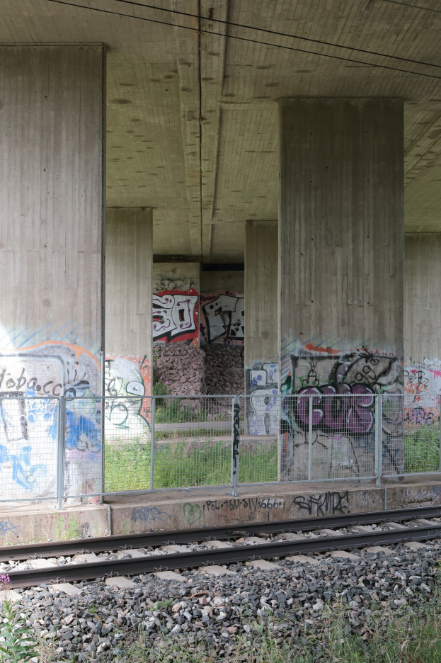 Eisenbahnschiene mit Graffiti, umgeben von Steinen, Gras, einem Zaun, Pfeilern, Drähten und einem Dach.