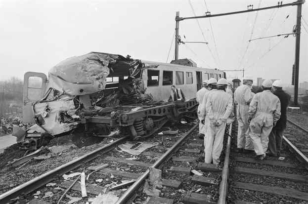 Schwarzes und weißes Bild einer Zugkollision auf den Schienen mit einer Gruppe von Helmträgern in der Nähe, umgeben von Strommästen, Drähten, Bäumen, Gebäuden und einem klaren Himmel.