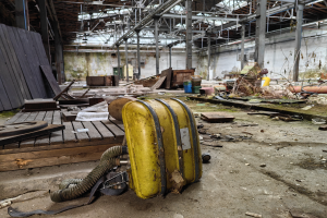 Ein gelber Koffer steht auf einem Holzfußboden in einem verlassenen Gebäude, umgeben von verstreuten Gegenständen wie Holzplanken und Papieren, mit einer Wand, Pfählen und einem Dach mit Metallstangen im Hintergrund.