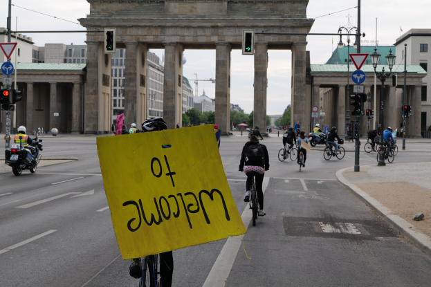 Eine Gruppe von Radfahrern in Helmen fährt eine Straße vor dem Brandenburger Tor in Berlin, Deutschland, entlang, wobei eine Person ein gelbes Schild hält, Lichtmasten, Verkehrsampeln, Gebäude, Bäume und einen klaren blauen Himmel im Hintergrund zu sehen sind.