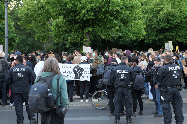 Eine große Gruppe von Menschen bei einer Black Lives Matter Demonstration in Berlin, einige halten Schilder, mit einem Fahrrad im Vordergrund und Bäumen und einem Pfahl im Hintergrund.