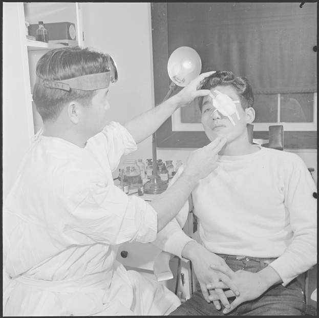 Schwarz-weiß-Foto eines Mannes, der von einem Arzt untersucht wird, mit medizinischer Ausrüstung und einem Fenster mit Vorhang im Hintergrund.