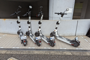 Drei schwarze und weiße E-Scooter mit Nummern und Text sind nebeneinander auf einem Gehweg vor einem Gebäude mit einer Glastür geparkt.