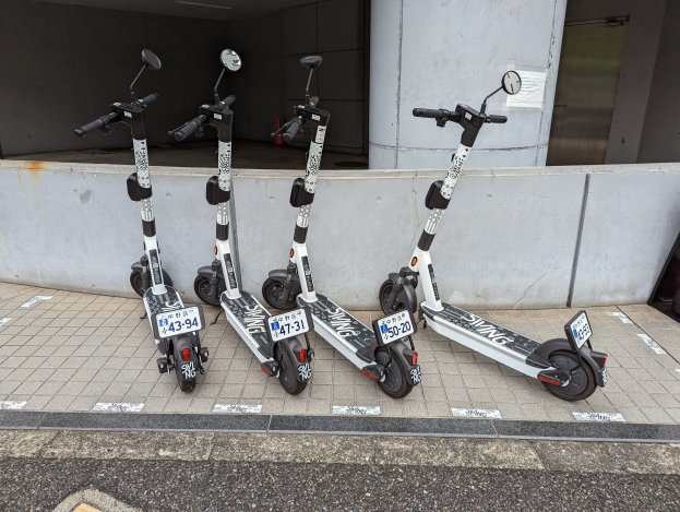 Drei schwarze und weiße E-Scooter mit Nummern und Text sind nebeneinander auf einem Gehweg vor einem Gebäude mit einer Glastür geparkt.