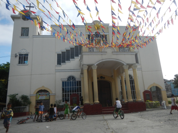 Eine Gruppe von Menschen, die vor einer weißen Kirche mit architektonischen Details wie Fenstern, Säulen, Bögen und einem Kreuzsymbol Fahrrad fahren, mit Pflanzen, einem Zaun, einer Fahne, Bäumen und einem bewölkten Himmel im Hintergrund und bunten Fahnen, die an einem Seil über dem Bild befestigt sind.