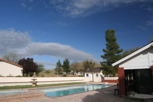 Ein Schwimmbad mit einem zentralen Brunnen, umgeben von grünem Gras, Bäumen und Gebäuden unter einem klaren blauen Himmel, mit einem Tisch und Stühlen auf der rechten Seite.