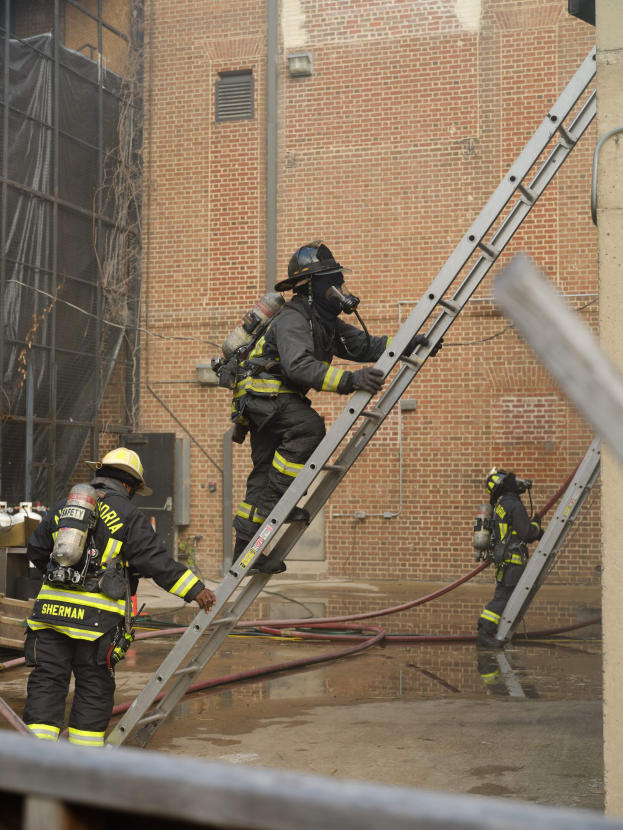 Feuerwehrleute in Helmen und Ausrüstung klettern an einer Leiter vor einem Backsteingebäude mit Rohren auf dem Boden und einer Metallstange unten, während im Hintergrund ein weiteres Gebäude mit Fenstern und einem Netz zu sehen ist.