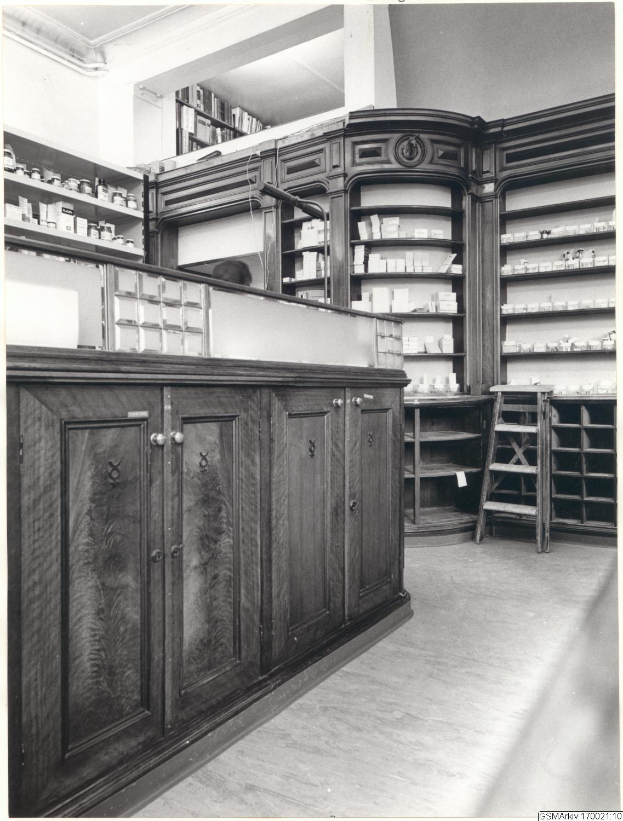 Schwarz-weiß-Foto eines vintage-Apothekeninneren mit einem Tresen, Schüben, einer Leiter und Regalen, die mit verschiedenen Gegenständen bestützt sind, mit Text oben.