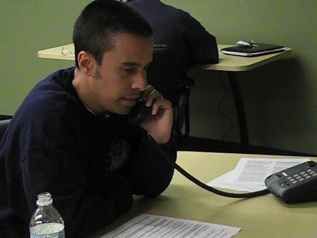 Mehrere Personen sitzen an einem Tisch mit einem Telefon, einer Einwegflasche und Kabeln. Eine Person spricht in das Telefon.