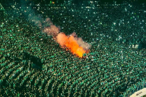 Luftaufnahme einer großen Menschenmenge in einem Stadion mit einem zentralen Feuer, das die Umgebung erhellt.