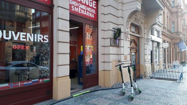 Eine Stadtstraße mit parkenden E-Scootern, gesäumt von Gebäuden mit Fenstern, Türen und Texttafeln, einem Geländer und Bäumen vor einem klaren blauen Himmel im Hintergrund.
