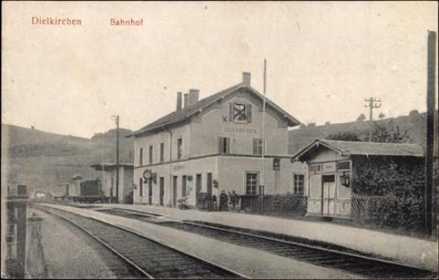 Altes Schwarz-Weiß-Foto eines Bahnhofs in Dielkirchen, Deutschland, mit einem Zug auf den Schienen, umgeben von Häusern, Pfählen, Bäumen, Pflanzen und Menschen, Hügel im Hintergrund und einem klaren Himmel.