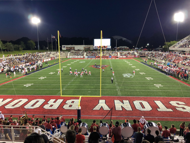 Fußballspiel in einem nächtlichen Stadion mit Spielern auf dem Feld, sitzenden Zuschauern, Musikern, einem Metallzaun, Lichtmasten, Fahnen, einem Anzeigebildschirm, Bäumen, Hügeln und einem sternenklaren Himmel.