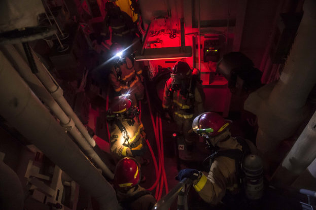Feuerwehrleute in Helmen und Sauerstoffflaschen stehen in einem schwach beleuchteten Raum mit Rohren auf beiden Seiten und bereiten sich auf einen Rettungseinsatz in der Nähe von Maschinen im Hintergrund vor.