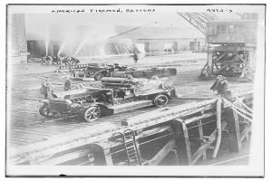 Ein Schwarz-Weiß-Foto eines Löschfahrzeugs auf einer Brücke mit einigen Menschen drumherum, eine Leiter führt hinauf zur Brücke, ein Gebäude im Hintergrund und Text oben auf dem Bild.