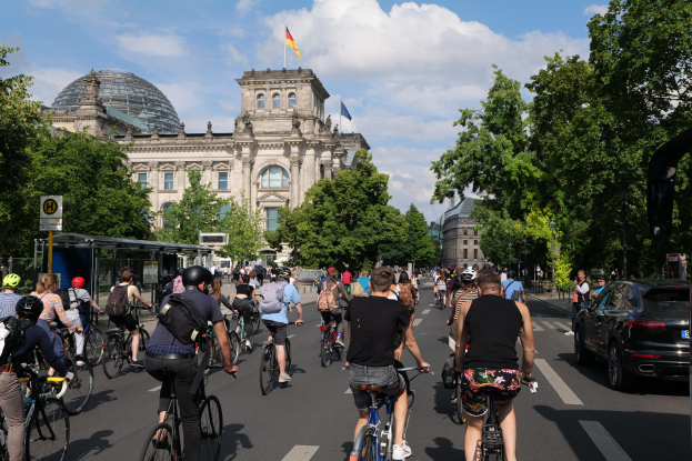 Eine Gruppe von Menschen, die auf Fahrrädern eine von Bäumen gesäumte Straße in Berlin, Deutschland, entlangfahren, mit Gebäuden und einer Bushaltestelle sichtbar, unter einem bewölkten Himmel und einer Flagge auf einem der Gebäude.
