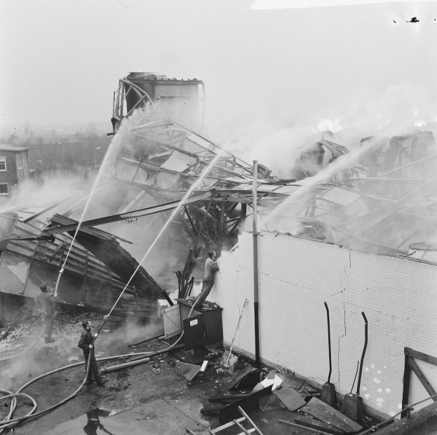 Schwarz-weiß-Foto von Feuerwehrleuten, die einen Dachbrand löschen, während Zuschauer von unten zusehen.