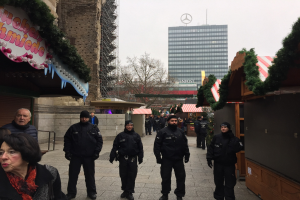 Polizisten in schwarzen Uniformen und Mützen stehen vor einem geschmückten Weihnachtsmarkt in Berlin, Deutschland, mit festlichen Dekorationen, Gebäuden, Bäumen und einem klaren blauen Himmel im Hintergrund.