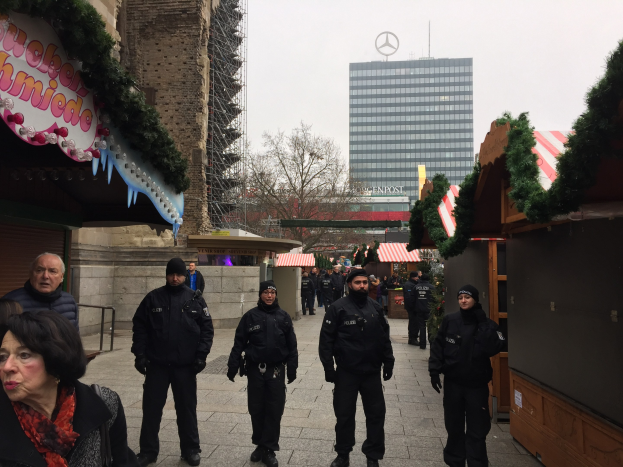 Polizisten in schwarzen Uniformen und Mützen stehen vor einem geschmückten Weihnachtsmarkt in Berlin, Deutschland, mit festlichen Dekorationen, Gebäuden, Bäumen und einem klaren blauen Himmel im Hintergrund.