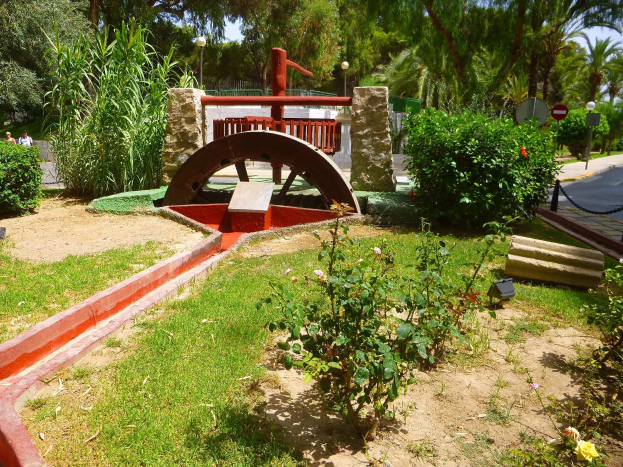 Minigolfanlage in einem Park mit Grünfläche, Straße, Passanten, Schildern, Bäumen und blauem Himmel.
