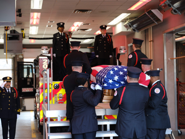 Gruppe uniformierter Männer, die einen Sarg tragen, der mit einer amerikanischen Flagge bedeckt ist, vor dem Hintergrund eines Feuerwehrautos und Deckenlampen.