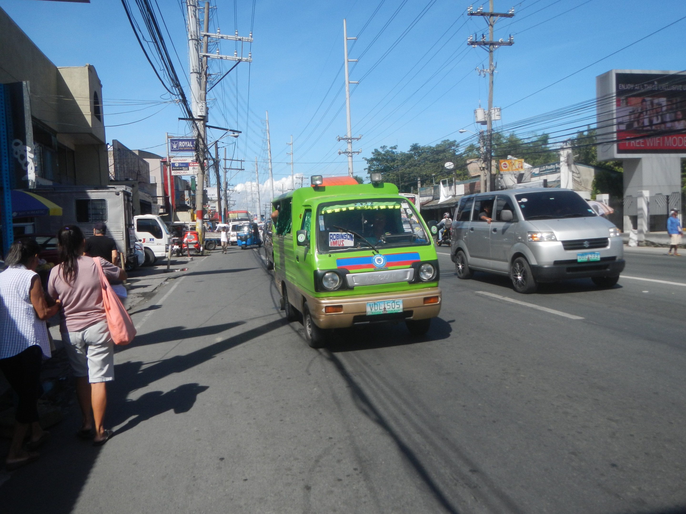 Ein grüner Lieferwagen fährt eine von hohen Gebäuden gesäumte Straße entlang, mit Passanten auf dem Bürgersteig, Strommasten und einem klaren Himmel im Hintergrund.
