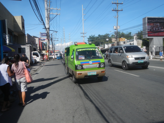 Ein grüner Lieferwagen fährt eine von hohen Gebäuden gesäumte Straße entlang, mit Passanten auf dem Bürgersteig, Strommasten und einem klaren Himmel im Hintergrund.